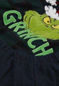 Grafika Zeleného Grinche na tmavém oblečení, s vystouplým písmem a měkkou texturou, s detailem na staženém elastickém pasu.