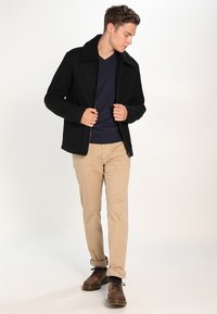 Schwarze Zip-Jacke mit einem Lammfellkragen, getragen über einem marineblauen Pullover, kombiniert mit beigen Hosen und braunen Schnürschuhen.