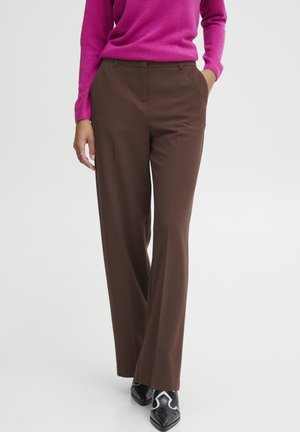 Person mit braunen, weit geschnittenen Hosen, magentafarbenem Pullover und spitzen schwarzen Schuhen mit weißen Details, stehend mit einer Hand in der Tasche.