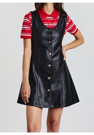 Femme portant une robe sans manches en simili cuir noir boutonnée, par-dessus une chemise à col rayée rouge et blanche, la main posée sur la hanche.