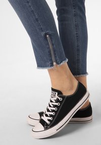 Svarta canvas-sneakers med vita gummisulor och tåhättor, i kombination med mörkblå skinny jeans med siddragkedjedetalj och fransad fåll.