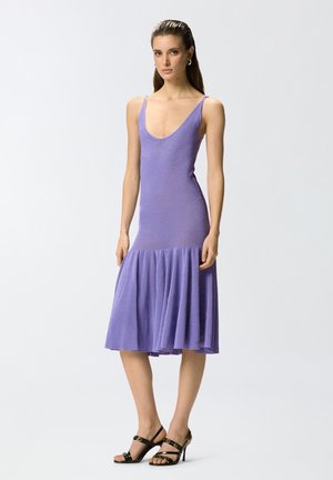 Mujer con un vestido sin mangas color lavanda, con escote redondo y bajo acampanado, combinado con sandalias negras de tacón alto y tiras.