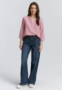 Chemise rose clair texturée à boutons, avec un décolleté en V et des manches trois-quarts, associée à un jean taille haute bleu foncé et des baskets blanches.