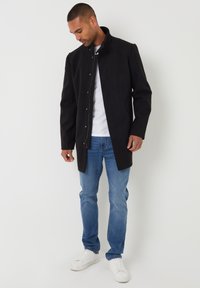 Sort, langærmet frakke med høj krave, tryklåslukning foran og sidelommer, parret med blå jeans og hvide sneakers.