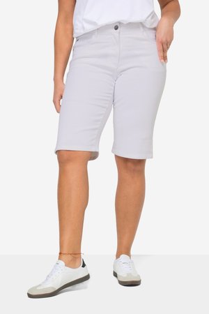 Person trägt hellgraue knielange Shorts, weiße Sneakers, weißes T-Shirt und ein rotes und gelbes Fußkettchen am linken Bein.