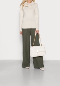Beige Rollkragenpullover, grüne weit geschnittene Hosen und eine weiße gesteppte Handtasche mit goldener Kette, ergänzt durch beigefarbene Blockabsatzschuhe.