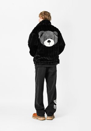 Persona de espaldas vistiendo una chaqueta de piel negra con una gran cara de oso gris en la espalda y pantalones negros con un oso más pequeño cerca del tobillo.