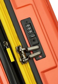 Esquina de maleta dura naranja con cremallera amarilla y ribete negro. Cuenta con un candado de combinación aprobado por la TSA y tiradores de cremallera metálicos.