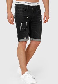 Crne traperice kratke hlače s izdistanciranim dijelovima, s odmjerenim rubovima i bijelim Calvin Klein logotipom na pojasu, u paru s bijelim tenisicama.