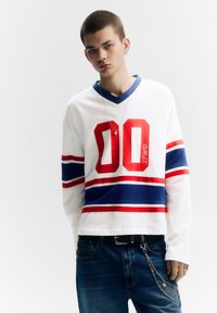 Fehér hosszú ujjú ing piros és kék csíkokkal, elöl feltűnő piros "00" számmal. Sötétkék farmerral és lánc részlettel párosítva.