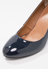 Pompe en cuir verni marine avec un bout rond et un design à découpes basses. L'intérieur présente une doublure en cuir lisse couleur sable. Fini brillant avec un éclat subtil.