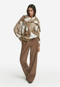 Beiger und weißer Strickpullover mit Diamantmuster und weiter Silhouette. Kombiniert mit braunen, weiten, gerippten Hosen und dunklen Schuhen.