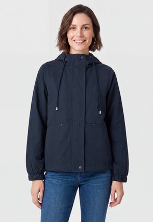 Donna con capelli corti castani sorride indossando una giacca blu navy con cappuccio e coulisse e jeans blu, in piedi davanti a uno sfondo chiaro e uniforme.