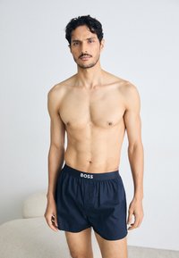 Temno modri boksarji z elastičnim pasom, ki vsebujejo logo "BOSS" in gumbasto zapiranje. Model stojí brez majice pred nevtralno ozadjem.