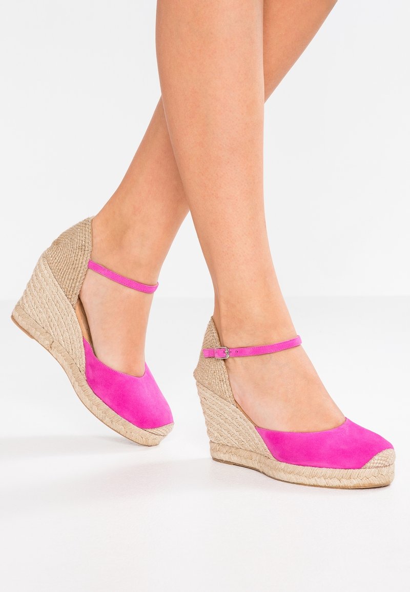 Jambes portant des espadrilles compensées en daim rose avec des brides à la cheville et des talons en jute tressée, posées sur une surface blanche.