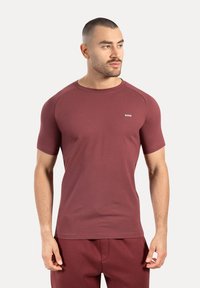 Man med kort hår och skägg som bär en åtsittande vinröd t-shirt och matchande byxor, ser åt sidan mot en enfärgad bakgrund.