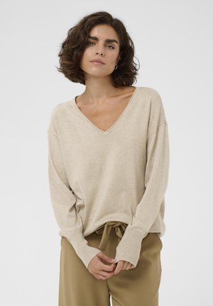 Donna con capelli corti e ricci, che indossa un maglione beige con scollo a V e pantaloni marrone chiaro con coulisse, in piedi davanti a uno sfondo bianco semplice.