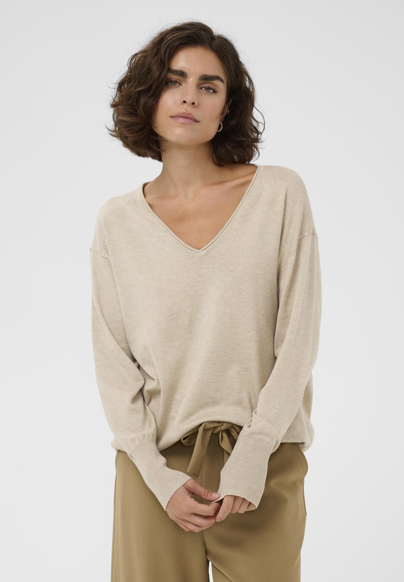 Femme aux cheveux courts bouclés portant un pull beige à col en V et un pantalon marron clair à cordon, debout devant un fond blanc uni.