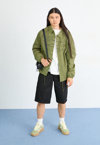 Chaqueta ligera verde con acentos negros, camiseta blanca debajo, pantalones cortos negros y zapatillas verdes con suelas texturizadas. Incluye bolso bandolera.