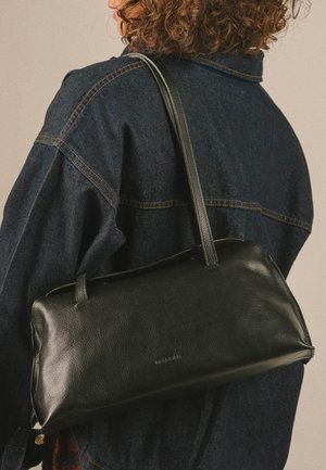 Persona con capelli ricci che indossa una giacca di jeans scura e porta una borsa a tracolla rettangolare in pelle nera con due cinturini.