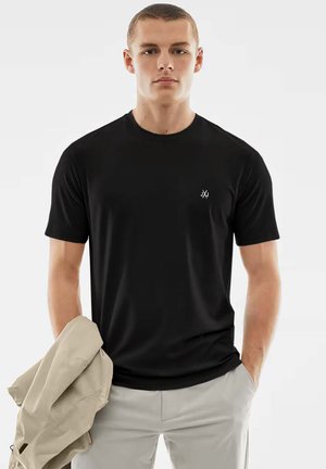 Black short-sleeve t-shirt made of smooth fabric, featuring a small white logo on the left chest. Model holds a light beige jacket.