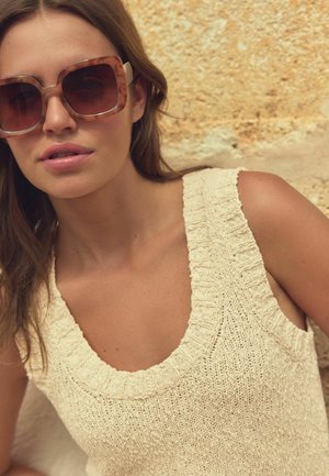Beige gestricktes Tanktop mit weitem Halsausschnitt und strukturierten Kanten. Das Modell trägt große, quadratische, gemusterte Sonnenbrillen vor einem neutralen Hintergrund.