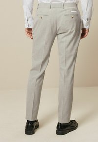 Pantalons sur mesure gris clair avec une finition texturée, deux poches arrière avec des accents dorés, et des jambes plissées, associés à des chaussures noires.