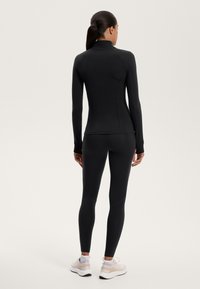 Top lungo aderente nero con maniche e leggings in tessuto elasticizzato. Presenta aperture per il pollice e cuciture piatte. Indossato con scarpe da ginnastica leggere.