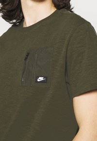 Tmavě zelené tričko s krátkým rukávem a texturovanou tkaninou, s kapsou na zip na hrudi a černou nášivkou s logem Nike.