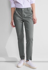Femme portant un pantalon gris ajusté avec une poche zippée et des baskets blanches, debout devant un fond clair uni.