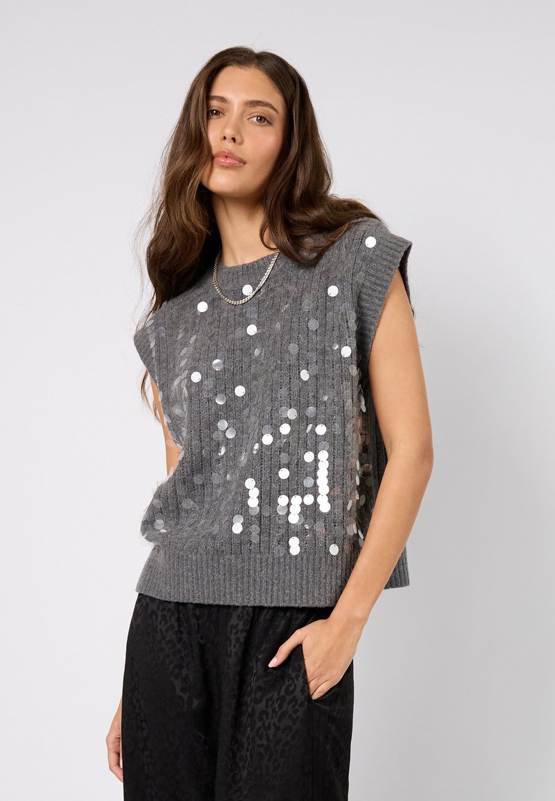 Pull sans manches gris en maille avec des sequins formant un motif sur le devant. La texture est douce avec des détails côtelés. Associé à un pantalon noir.