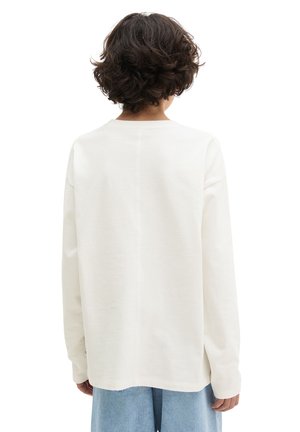 Person mit kurzen lockigen braunen Haaren, die ein locker sitzendes weißes Langarmshirt und hellblaue Jeans trägt, von hinten gesehen.