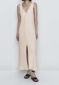 Strick, ärmelloses V-Ausschnittkleid in hellbeige mit einem Schlitz vorne, kombiniert mit schwarzen Riemchensandalen. Glatte Textur und knöchellanges Design.