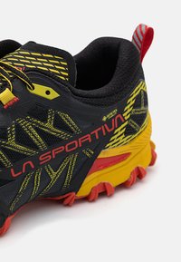 Črne in rumene tekaške čevlje za trail z teksturirano mrežasto zgornjico, ojačanimi stranicami, rdečimi akcenti in trpežnim, večbarvnim gumijastim podplatom.
