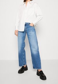 Bílá košile na knoflíky s kapsou, kombinovaná s volnými světle modrými džíny. Džíny mají vybledlý vzhled a jsou stylizovány s černými platformovými botami.