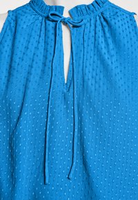 Top azul sem mangas com um padrão texturizado de bolinhas, decote franzido e uma abertura frontal em forma de chave com laços. Tecido suave com um design fluído.