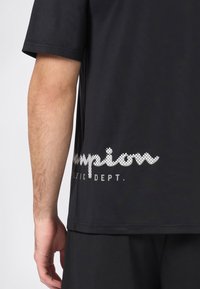Acercamiento del brazo de una persona y el torso lateral vistiendo una camiseta negra con el texto blanco punteado "Champion Athletic Dept." cerca del dobladillo.
