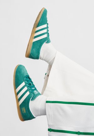 Türkisfarbene Veloursleder-Sneaker mit weißen Streifen und Gummisohle. Mit Schnürsenkeln und Markendetails; getragen mit weißen Socken und passenden Hosen.