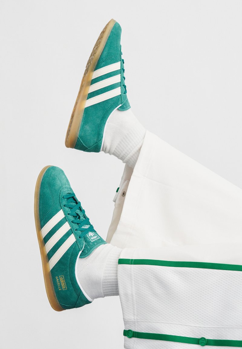 Sapatilhas de camurça azul-petróleo com riscas brancas e sola de borracha castanha. Apresentam atacadores e detalhes de marca; usadas com meias brancas e calças a condizer.