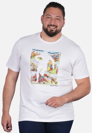 Homme barbu souriant, portant un t-shirt blanc avec des scènes de rugby colorées de style bande dessinée et une grande silhouette blanche de joueur de rugby.