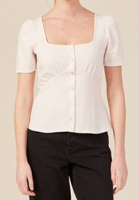 Blusa beige chiaro a maniche corte con scollo a quadrato, tessuto testurizzato e chiusura frontale con bottoni. Presenta dettagli arricciati in vita.