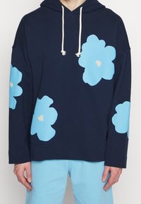 Felpa con cappuccio blu navy con grandi motivi floreali azzurri, dotata di cappuccio con coulisse e polsini a costine. Abbinata a pantaloni azzurri.