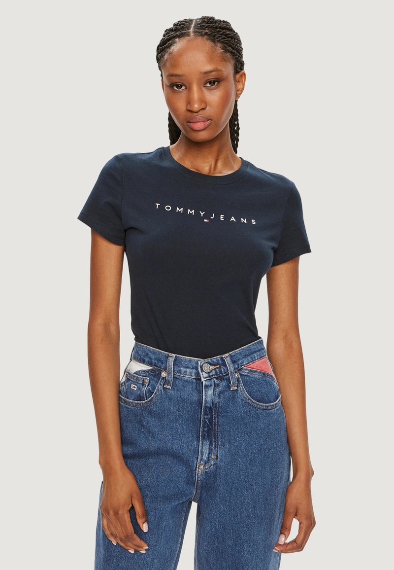 Femme portant un t-shirt bleu marine "TOMMY JEANS" rentré dans un jean taille haute bleu avec des patchs en tissu coloré sur les poches.