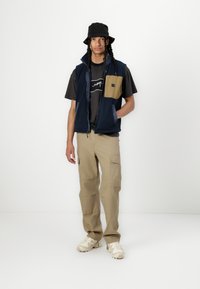 Marinevest med beige accenter, sort t-shirt med hvidt design, beige cargo bukser og cremede sneakers. Sort bucket hat.