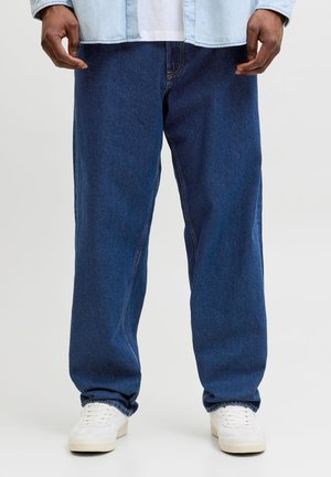 Άνδρας που φοράει άνετα σκούρα μπλε τζιν, λευκά αθλητικά παπούτσια και ανοιχτό μπλε πουκάμισο από τζιν, με τα χέρια χαλαρά στα πλευρά.