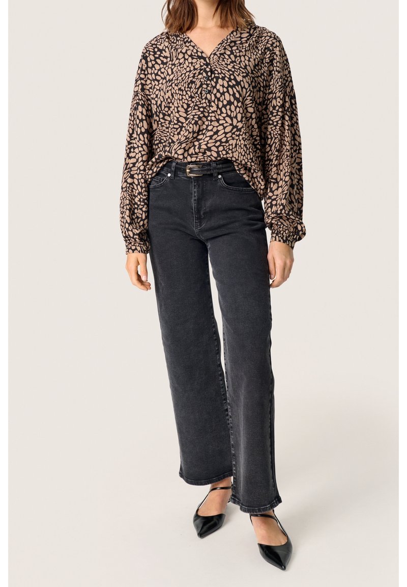 Femme portant un chemisier à manches longues à motifs marron, rentré dans un jean noir taille haute à jambes larges, avec des chaussures noires pointues.