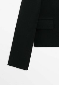Manche d'un manteau en laine noire et partie du corps avec un détail de poche à rabat sur fond blanc.