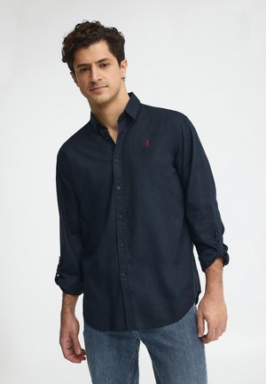 Giovane uomo con capelli ricci che indossa una camicia blu navy con bottoni e maniche rimboccate e jeans blu, in piedi davanti a uno sfondo semplice.