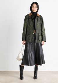 Ελιάς πράσινο αδιάβροχο μπουφάν με καφέ κοτλέ γιακά, δύο μπροστινές τσέπες, συνδυασμένο με μαύρη δερμάτινη φούστα και μαύρες μπότες  μέχρι το γόνατο.