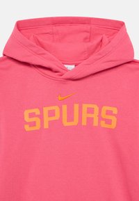 Różowa bluza z kapturem ze sznurkiem, z napisem "SPURS" w wyraźnych pomarańczowych literach oraz małym pomarańczowym logo Nike swoosh nad napisem. Miękka dzianina dżersejowa.
